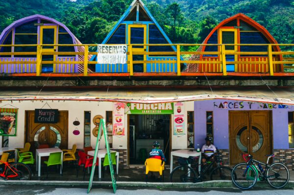 Colorful buildings with triangular roofs overlook a bustling street in Villa Restrepo, Tolima, showcasing vibrant outdoor seating and bicycles.