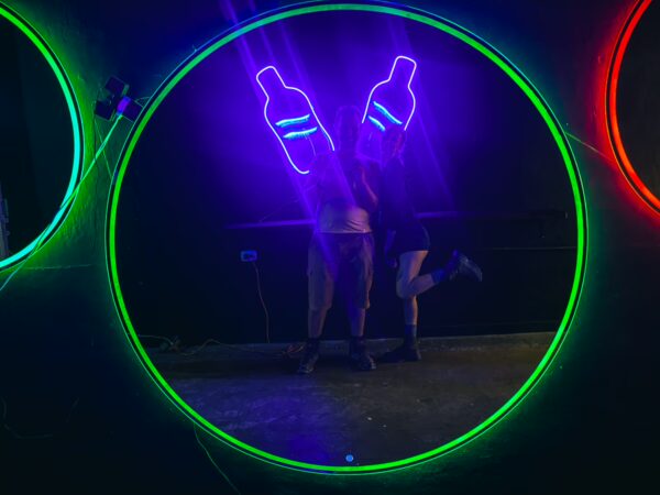Brightly lit backdrop with neon bottle props and two friends playfully posing together in a colorful setting in Quindio, Colombia.