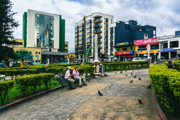 Colorful buildings surround a lush green park in Santa Rosa de Cabal, with people sitting on benches and pigeons scattered along the pathway.