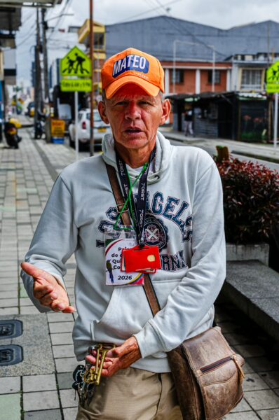 A man wearing a bright orange cap stands on a sidewalk in Quindio, Colombia, gesturing with a small brass instrument in hand.
