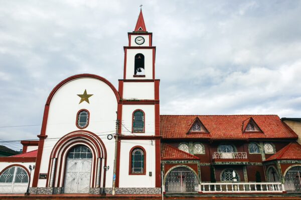 Colorful church with red accents and a clock tower in Medellia, Milagrosa, Murillo, Tolima, Colombia under a cloudy sky.