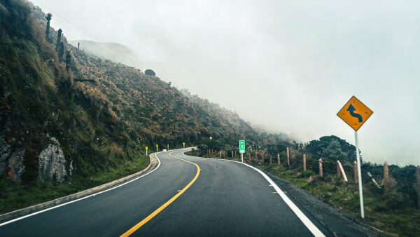 Winding road leads through foggy landscape near Nevado del Ruiz, showcasing lush greenery and a caution curve sign in Tolima, Colombia.