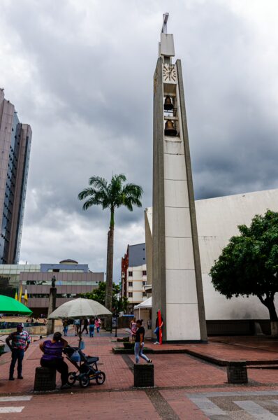 Prominent clock tower stands in Plaza de Bolívar, surrounded by trees and visitors, under a dramatic cloudy sky in Armenia, Quindío, Colombia.