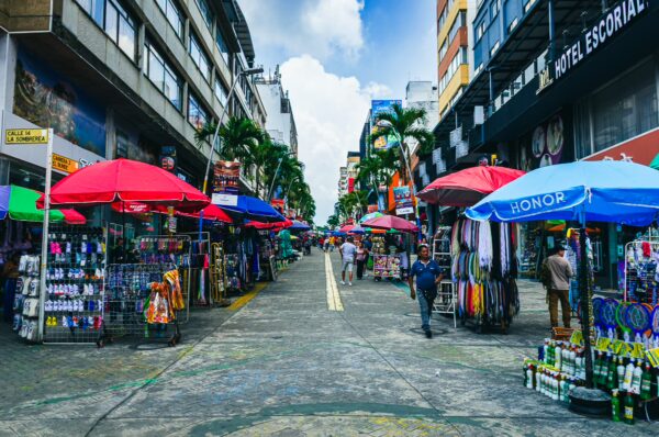 Colorful market stalls with red and blue umbrellas line the street in Ibagué, Tolima, Colombia, bustling with shoppers and vendors.