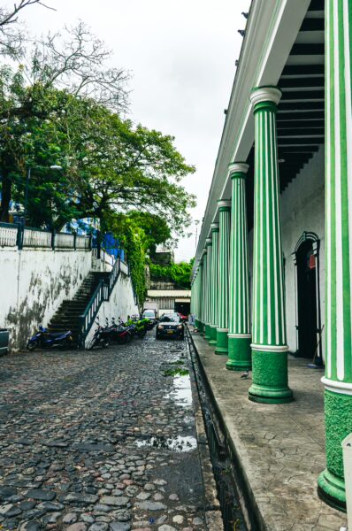 Green columns line a cobblestone street in Plaza de Mercado, Honda, Tolima, Colombia, featuring lush trees and parked vehicles.
