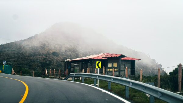 Curving road with a guardrail leads to a rustic shelter amidst misty hills on Nevado del Ruiz, Tolima, Colombia.