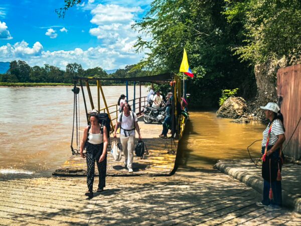 Travelers disembark from a small ferry at the riverside in Villavieja, Huila, Colombia, surrounded by lush greenery and blue skies.