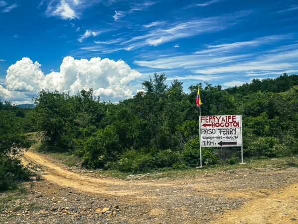 A dirt road leading towards a sign for Ferretería La Viejita, surrounded by green vegetation and blue sky in Villavieja, Huila, Colombia.