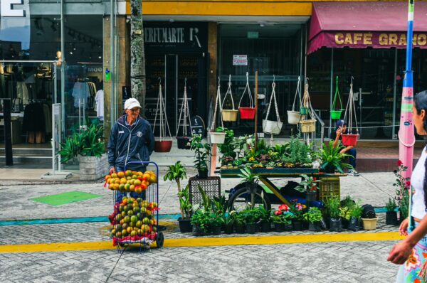 Vibrant fruit vendor cart filled with fresh produce, surrounded by green plants and shops along Carrera 3 in Ibagué, Tolima, Colombia.