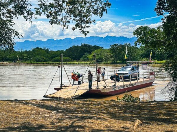 A ferry carrying a truck crosses the river in Villavieja, Huila, with lush greenery and mountains in the background.