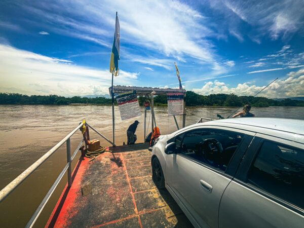 Ferry crossing the river in Villa Vieja, Huila, Colombia, with a car on board and flags fluttering in the sunny sky.