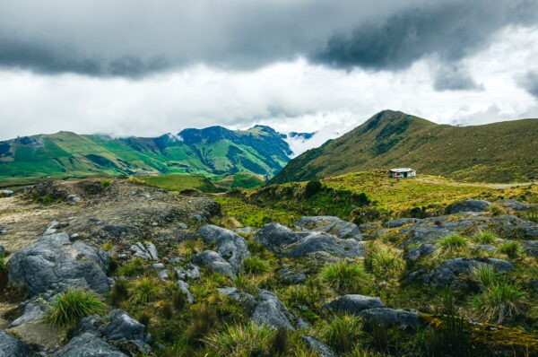 Lush green hills and rocky terrain under a cloudy sky near Nevado del Ruiz, Tolima, Colombia, with a small structure in the distance.