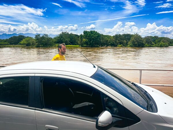 A person stands on the roof of a white car, overlooking the muddy river against a backdrop of lush greenery in Villavieja, Huila, Colombia.