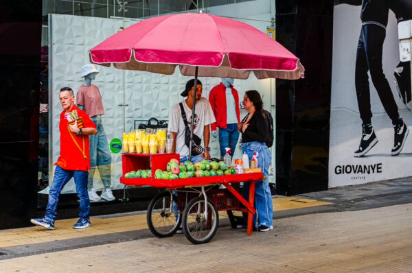 Bright pink umbrella shades a fruit cart with locals chatting outside a shopping area in Armenia, Quindío, Colombia.