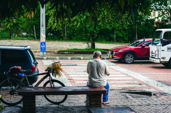 A man sits on a wooden bench near a bicycle, surrounded by greenery and parked cars in Honda, Tolima, Colombia.