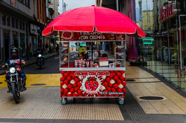 Brightly colored food cart selling ice cream, adorned with red fruit designs, in the lively streets of Armenia, Quindío, Colombia.