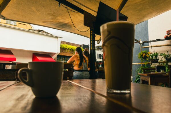 Warm coffee and a frothy drink sit on a wooden table at a café in Santa Rosa de Cabal, Risaralda, Colombia, with vibrant greenery nearby.