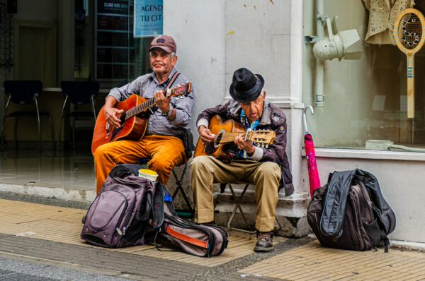 Two musicians play guitars on a street in Armenia, Quindío, Colombia, surrounded by backpacks and a lively urban backdrop.