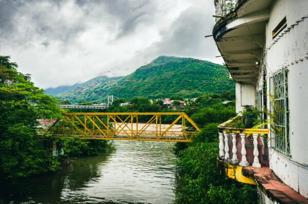 Yellow bridge spans the river, surrounded by lush green hills and a cloudy sky in Tolima, Colombia.