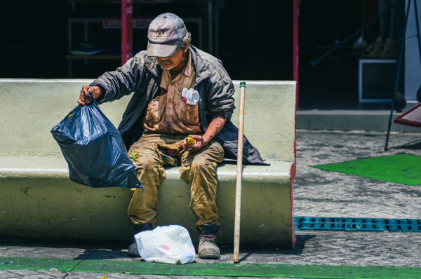 A man in worn clothing sorts through a garbage bag while seated on a green bench in Ibagué, Tolima, Colombia.