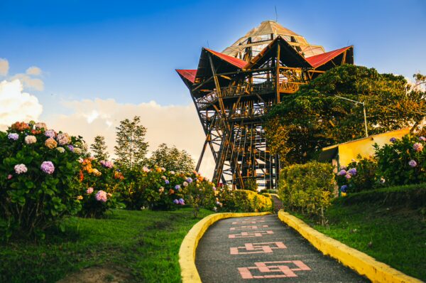 Colorful flowers line the path leading to the Mirador Colina Iluminada tower in Filandia, Quindio, Colombia, against a bright sky.