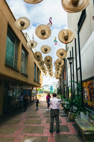 Colorful sombreros hang overhead along a vibrant street in Plaza Dario Echandía, Ibagué, Tolima, Colombia, with people strolling below.