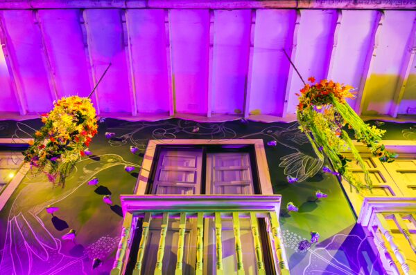 Colorful floral arrangements hang from a vibrant, purple-lit balcony in Santa Rosa de Cabal, Risaralda, Colombia.