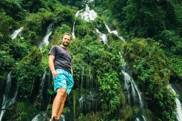 Vibrant waterfalls cascade down lush greenery, with a traveler in swim shorts enjoying the natural beauty at Termales Rosa in Risaralda, Colombia.