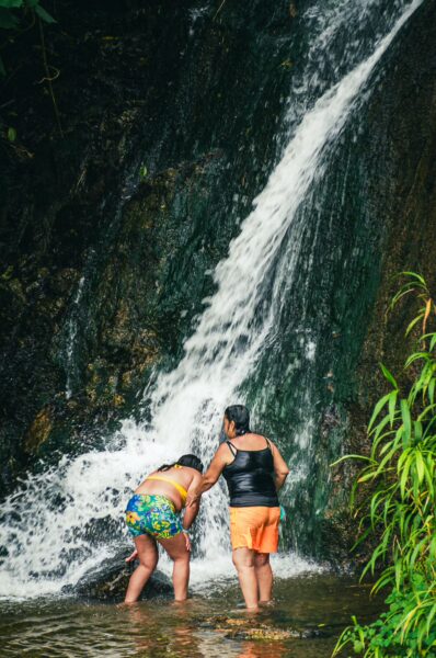 Two people explore the base of a waterfall surrounded by lush greenery at Termales Rosa in Risaralda, Colombia.