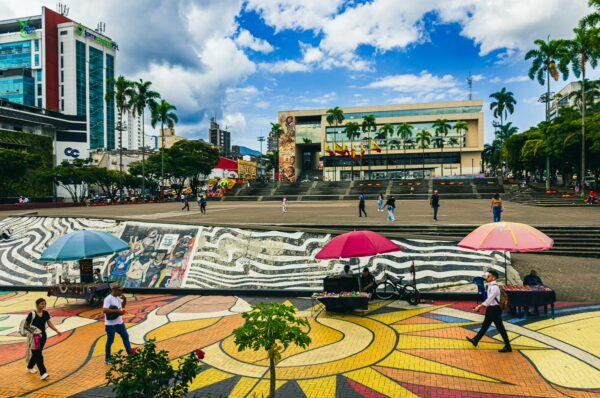 Colorful murals and umbrellas decorate the lively Plaza Ciudad Victoria in Pereira, Risaralda, under a vibrant blue sky.
