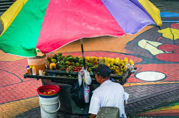 Vibrant fruit vendor under a colorful umbrella in Plaza Ciudad Victoria, Pereira, showcasing fresh produce and lively street art.