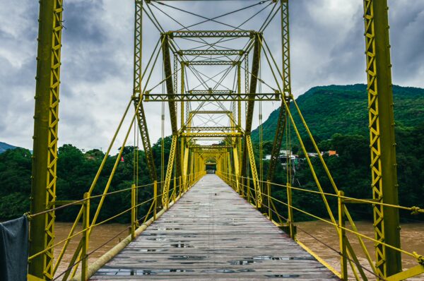 Yellow metal suspension bridge spans a river, framed by lush green hills under a cloudy sky in Honda, Tolima, Colombia.