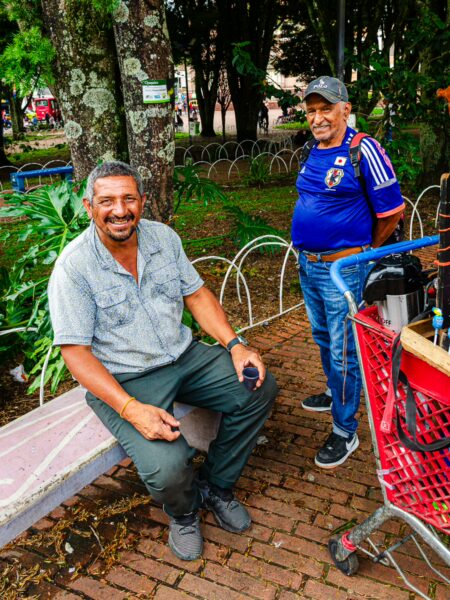 Two smiling men sit on a bench in a park in Armenia, Quindío, surrounded by lush greenery and a brick walkway.