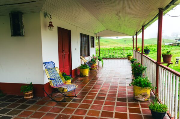 Colorful rocking chairs and potted plants line a covered porch at a rustic house in Murillo, Tolima, Colombia, surrounded by lush greenery.