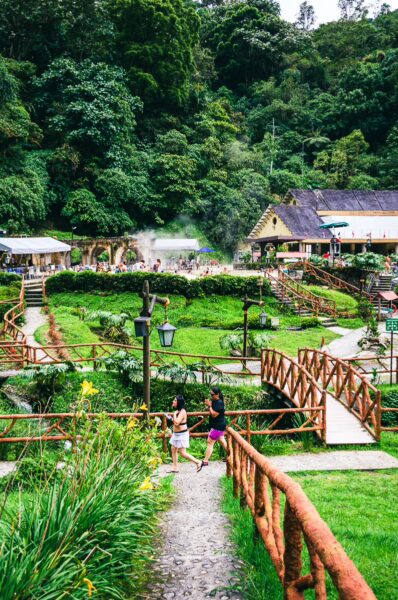Visitors walk along a scenic path through lush greenery at Termales de Rosa, Risaralda, Colombia, with wooden bridges and hot springs.