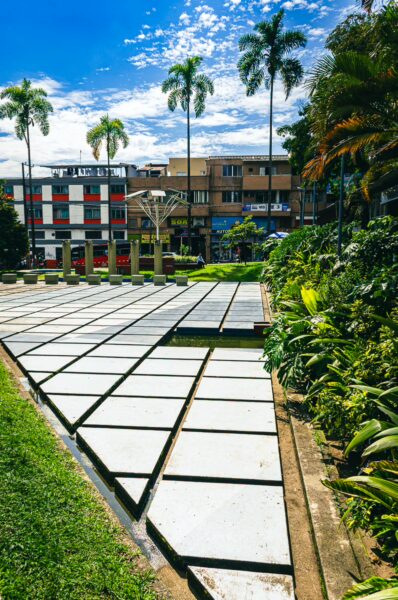 Modern plaza layout with geometric pathways, lush greenery, and palm trees under a bright sky in Ciudad Victoria, Pereira, Risaralda.