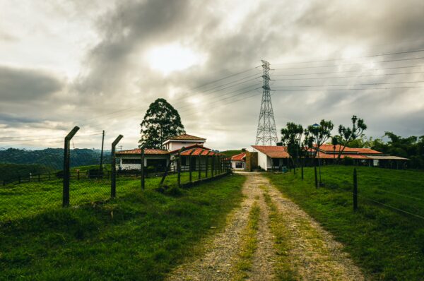 Dusty path leads to a rustic building with a red roof, surrounded by green hills under a cloudy sky in Quindío, Colombia.