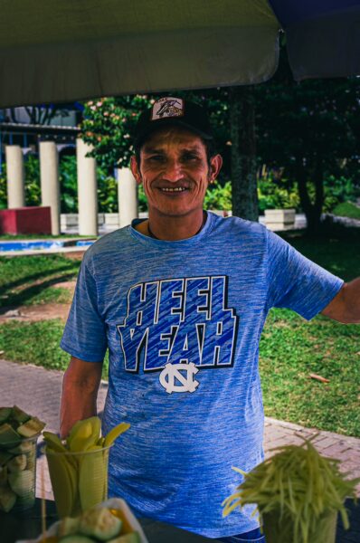 Proud vendor in a blue shirt smiles while offering fresh fruits at a vibrant street market in Pereira, Risaralda, Colombia.