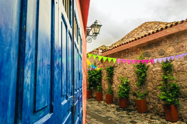 Vibrant blue doors and colorful banners adorn Calle de las Trampas Honda, featuring lush greenery and rustic stone walls in Tolima, Colombia.