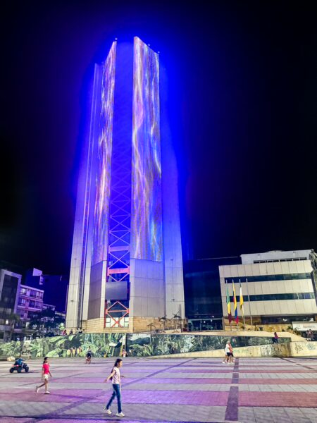 Vibrant tower illuminated in blue, surrounded by pedestrians in a lively plaza in Armenia, Quindío, Colombia.