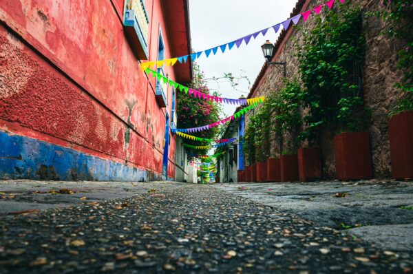 Colorful flags drape across the cobblestone street of Calle de las Trampas Honda, lined with vibrant buildings and greenery in Tolima, Colombia.