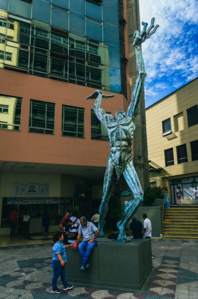El Gran Hombre statue stands tall in Pereira, Risaralda, Colombia, surrounded by visitors and modern architecture under a blue sky.