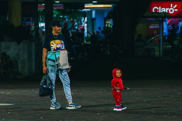 A man carrying a bag stands next to a smiling child in a red outfit, set against the vibrant backdrop of Plaza de Bolívar, Pereira.