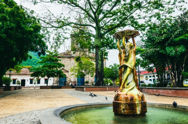 Golden fountain sculpture surrounded by lush greenery in Jose Leon Armero Park, Honda, Tolima, Colombia. Historic buildings in background.