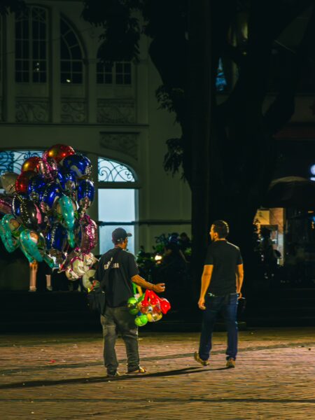 Colorful balloons are held by a vendor in Plaza de Bolívar, with a visitor walking towards him in Pereira, Risaralda, Colombia.