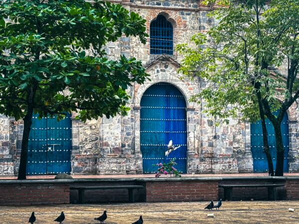 Vibrant blue doors stand out against the stone wall of Jose Leon Armero Park in Honda, Tolima, with lush trees and pigeons nearby.