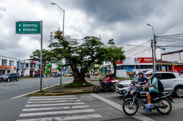 Busy street scene in Quindío, Colombia, featuring motorcycles, pedestrians, and a large tree, with hospital signage visible.