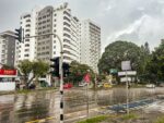 Rain-soaked street scene in Quindio, Colombia, featuring modern buildings, traffic lights, and lush greenery under a cloudy sky.
