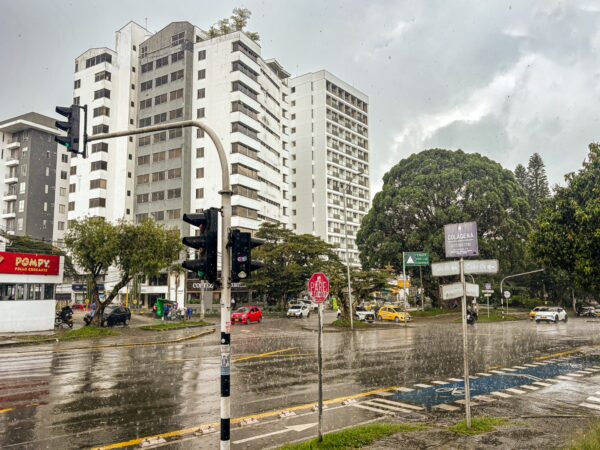 Rain-soaked street scene in Quindio, Colombia, featuring modern buildings, traffic lights, and lush greenery under a cloudy sky.