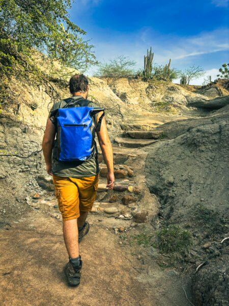 Hiker in a blue backpack navigating rocky terrain with cacti under a bright sky in Tatacoa Desert, Huila, Colombia.